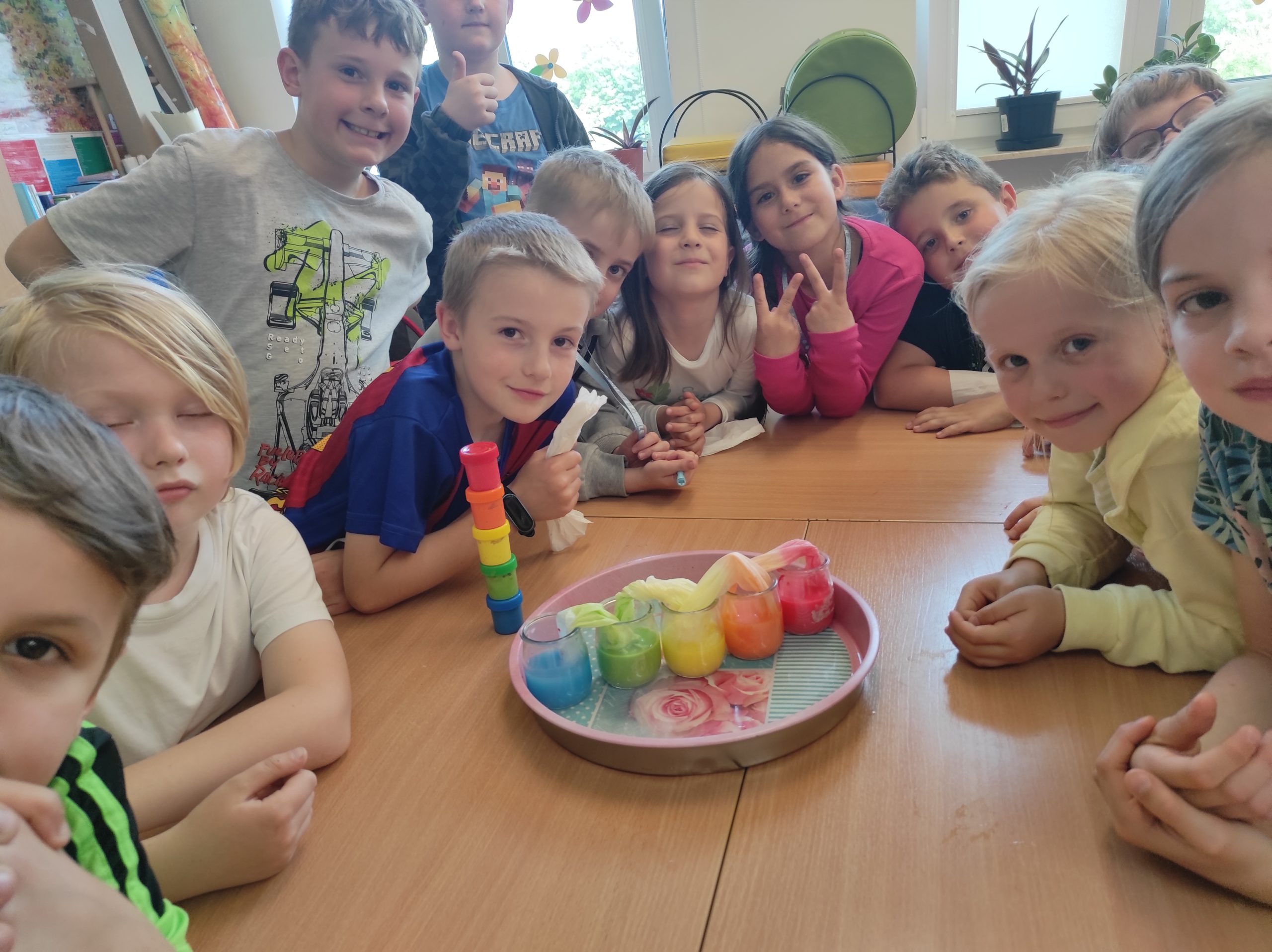 13 dzieci siedzi wokoło złączonych stołów. Na środku leży taca, a w niej 5 słoiczków. W każdym z nich jest inny kolor płynu. Słoiczki są połączone ze sobą kolorowymi papierami. W tle elementy sali.