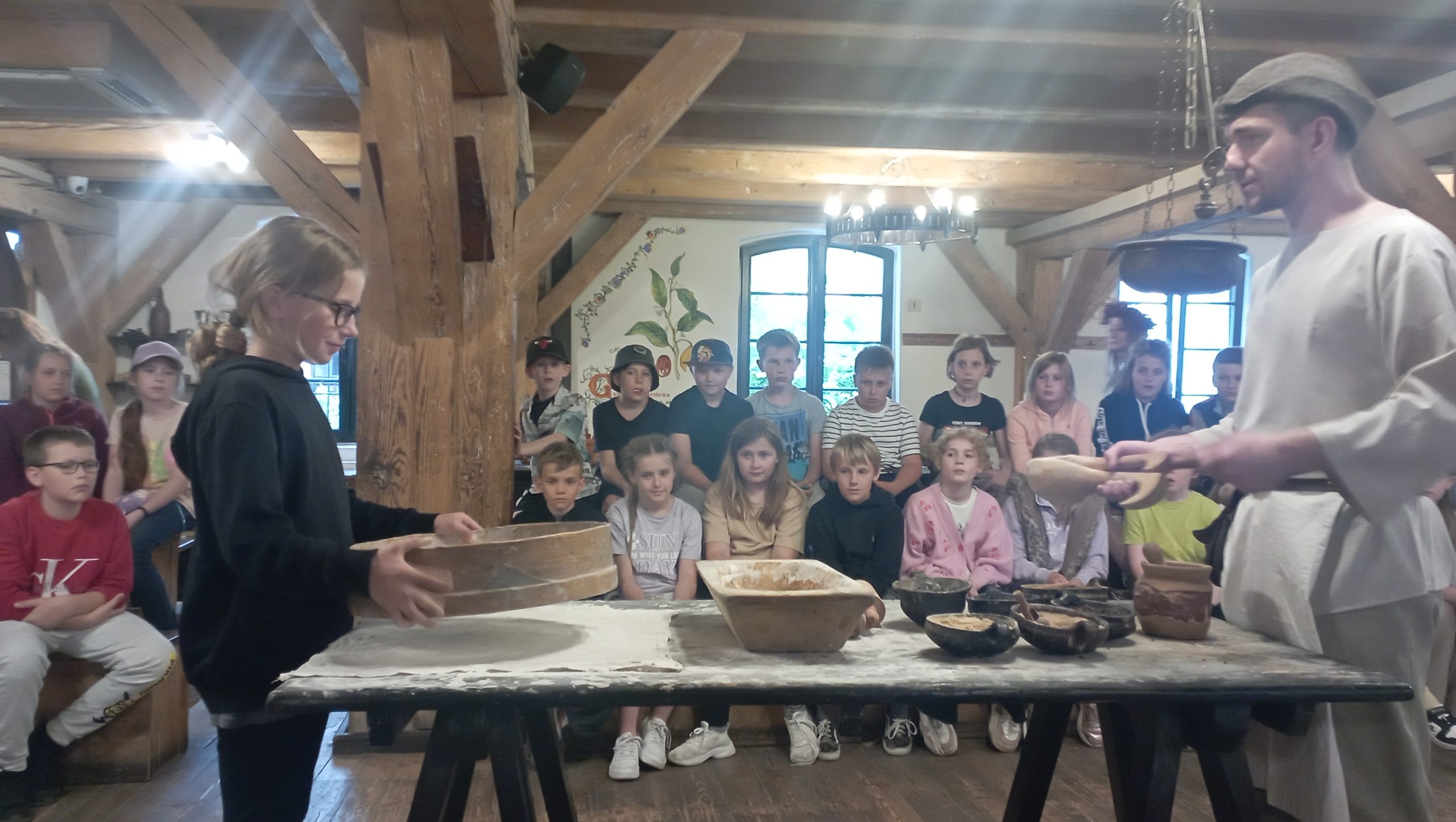 W Muzeum Piernika dziewczynka przesiewa sitem mąkę. W tle siedzące dzieci.