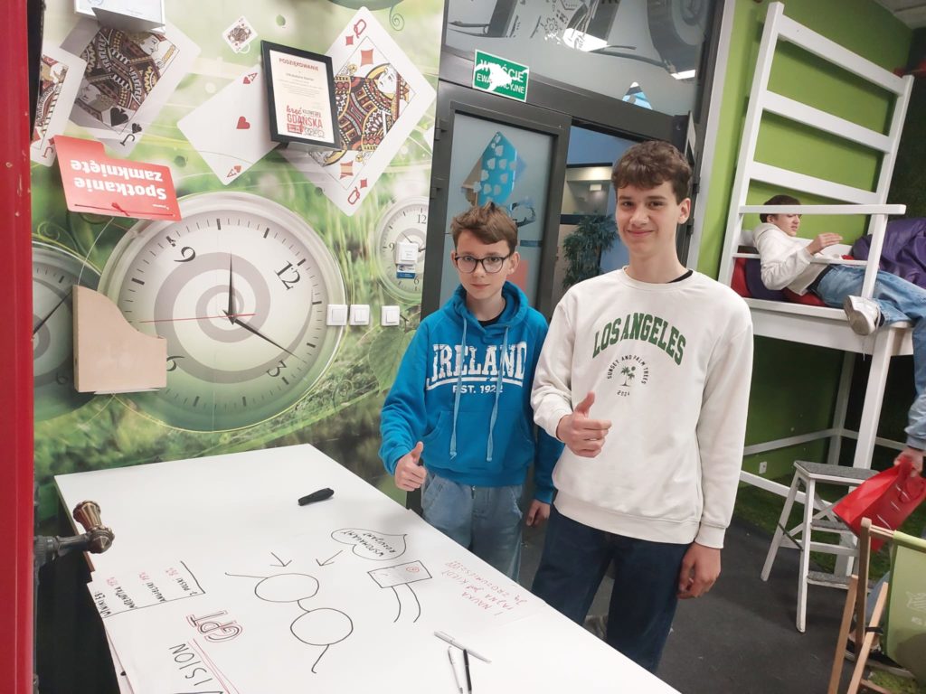 Dwóch nastolatków stojących przy białym stole z dużym arkuszem papieru, na którym widnie-ją rysunki i schematy. Obaj chłopcy mają kciuki uniesione w geście aprobaty. Jeden z nich ma na sobie niebieską bluzę z napisem „Ireland”, a drugi biały sweter z napisem „Los Angeles”. W tle widać kolorowe, inspirujące wnętrze z dużymi grafikami zegarów i kart do gry na ścia-nach, a także innych uczniów zaangażowanych w pracę lub odpoczywających na podestach.