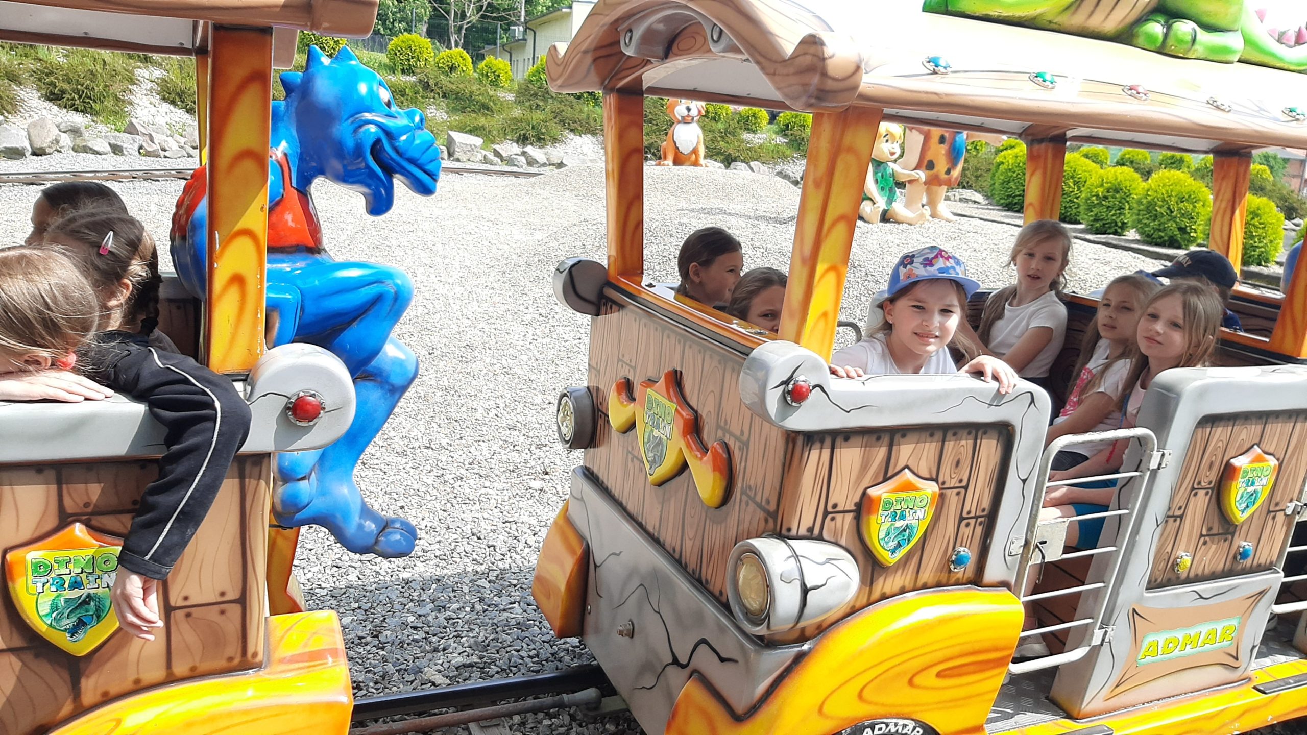 .Zdjęcie w plenerze. Fragmenty dwóch wagoników stylizowanych na pocią jaskiniowców z tarczami DINO TRAIN. W środku siedzą dzieci. Na tyle pierwszego wagonu siedzi figura dinozaura. W tle kamienista skarpa z zielenią.