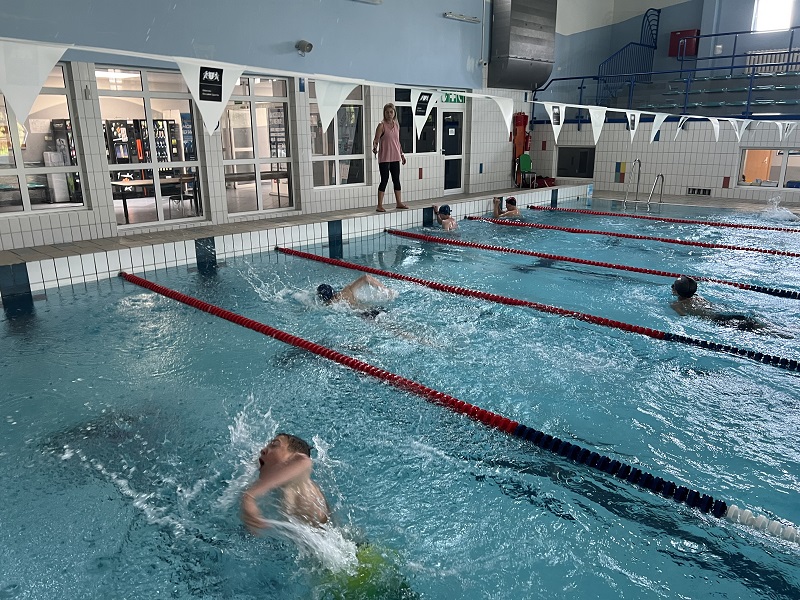 Pięciu uczniów kończy wyścig pływacki, dwóch po prawej dopłynęło , a trzech z lewej pokonuje ostatnie metry. Na brzegu stoi pani, mierząca czasy