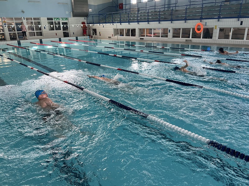 Pięciu uczniów płynie kraulem w wyścigu. W tle pani mierząca czasy.