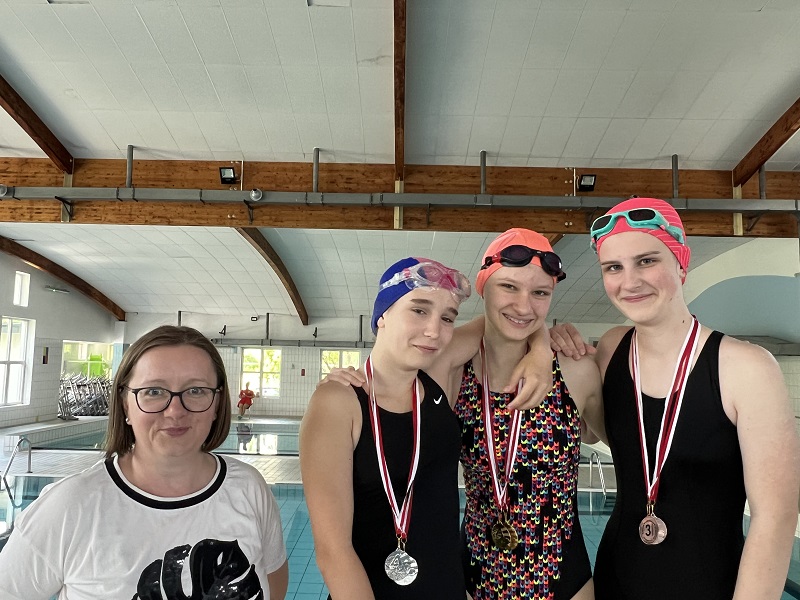 Trzy uczennice z medalami na szyi pozują do zdjęcia z panią stojącą z lewej strony