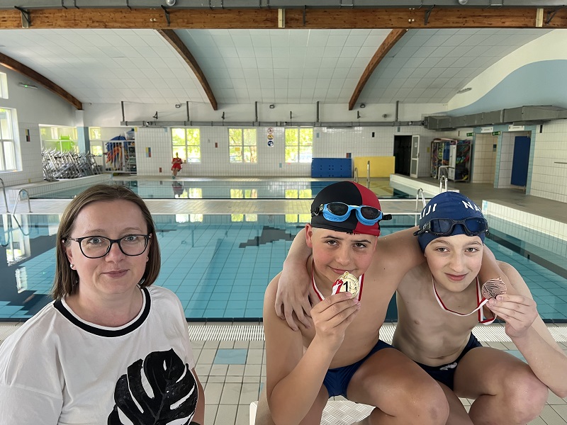 Dwóch uczniów kucających na podium z medalami w dłoni pozuje do zdjęcia z panią stojącą z lewej