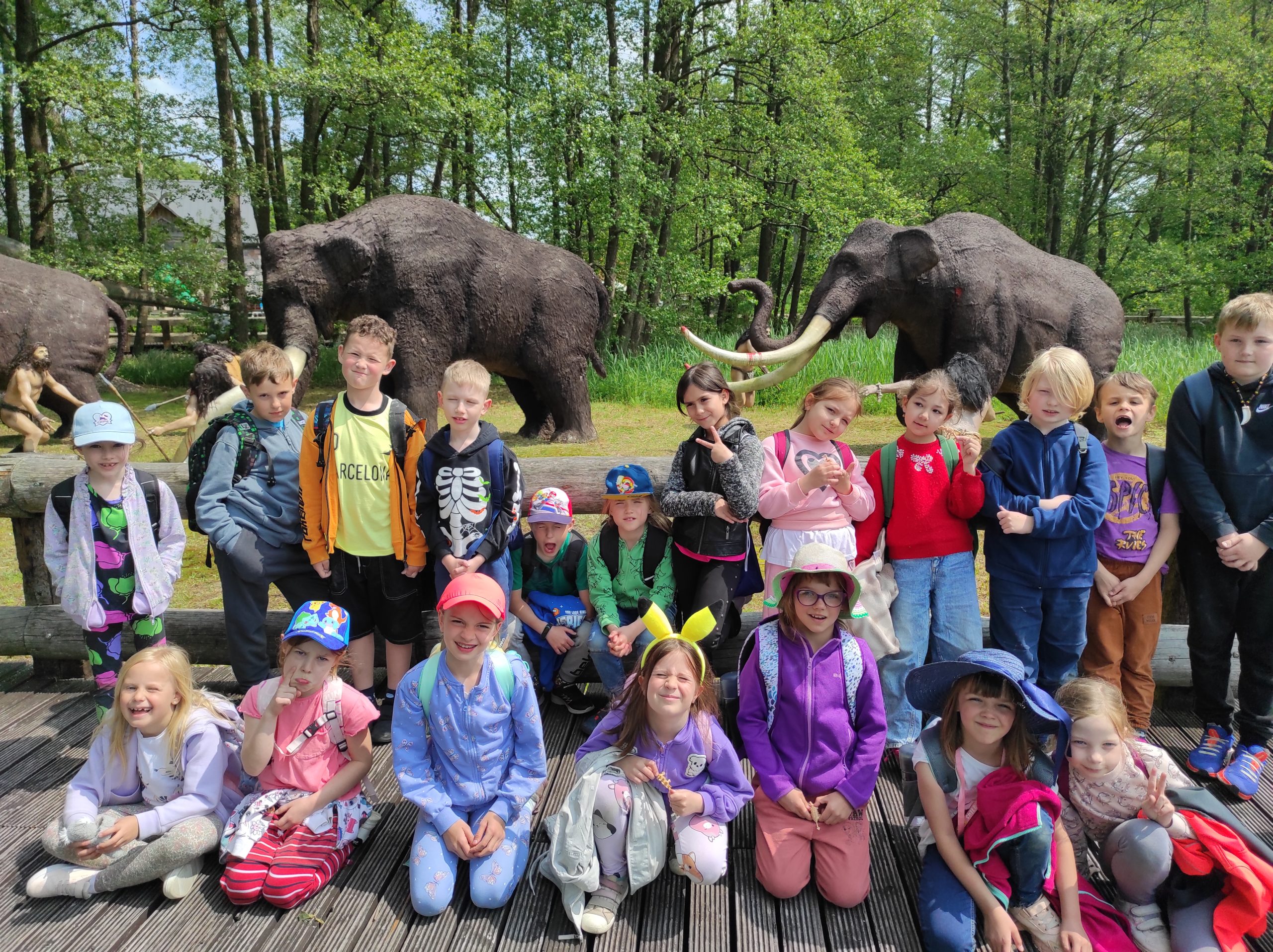 Zdjęcie w plenerze. Dzieci ustawione w 2 rzędy, jeden stoi, drugi siedzi. W tle zieleń oraz dwie duże  makiety mamutów.