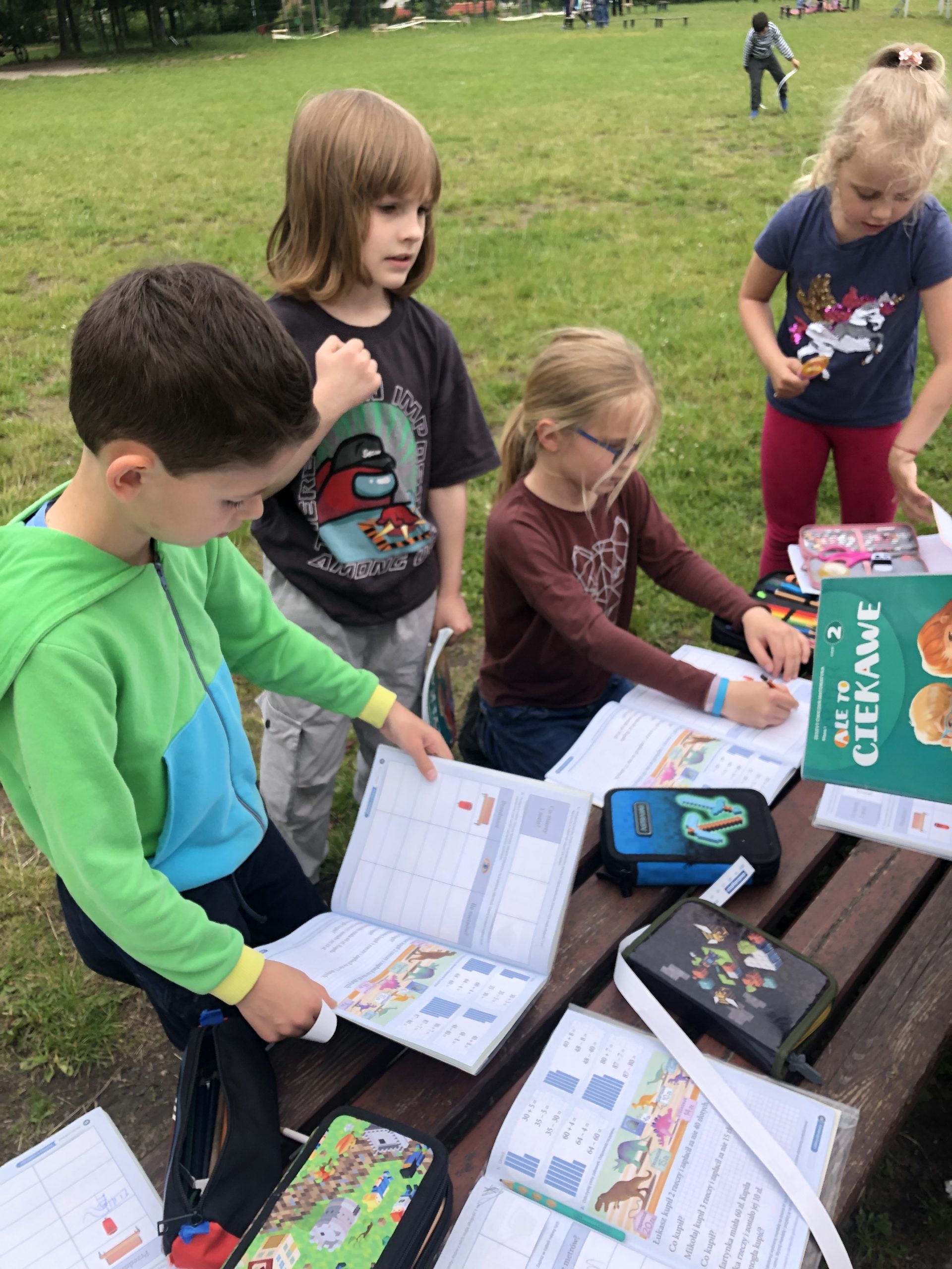 Czwórka uczniów zebrana przy ławce na boisku szkolnym notuje w ćwiczeniach wyniki pomiarów.