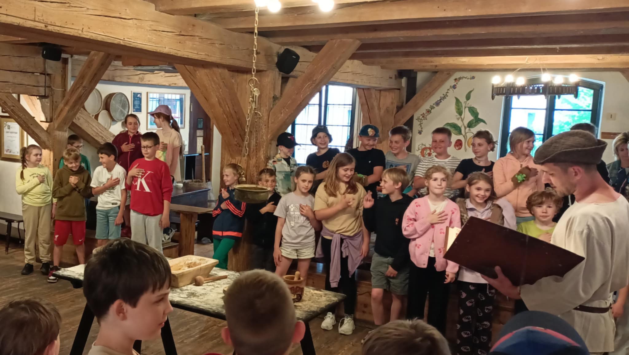 Uczniowie podczas warsztatów w Muzeum Piernika składają przysięgę. Wszyscy stoją i trzymają dłoń na sercu. Na środku sali jest stół, na którym leżą produkty do robienia ciasta piernikowego.