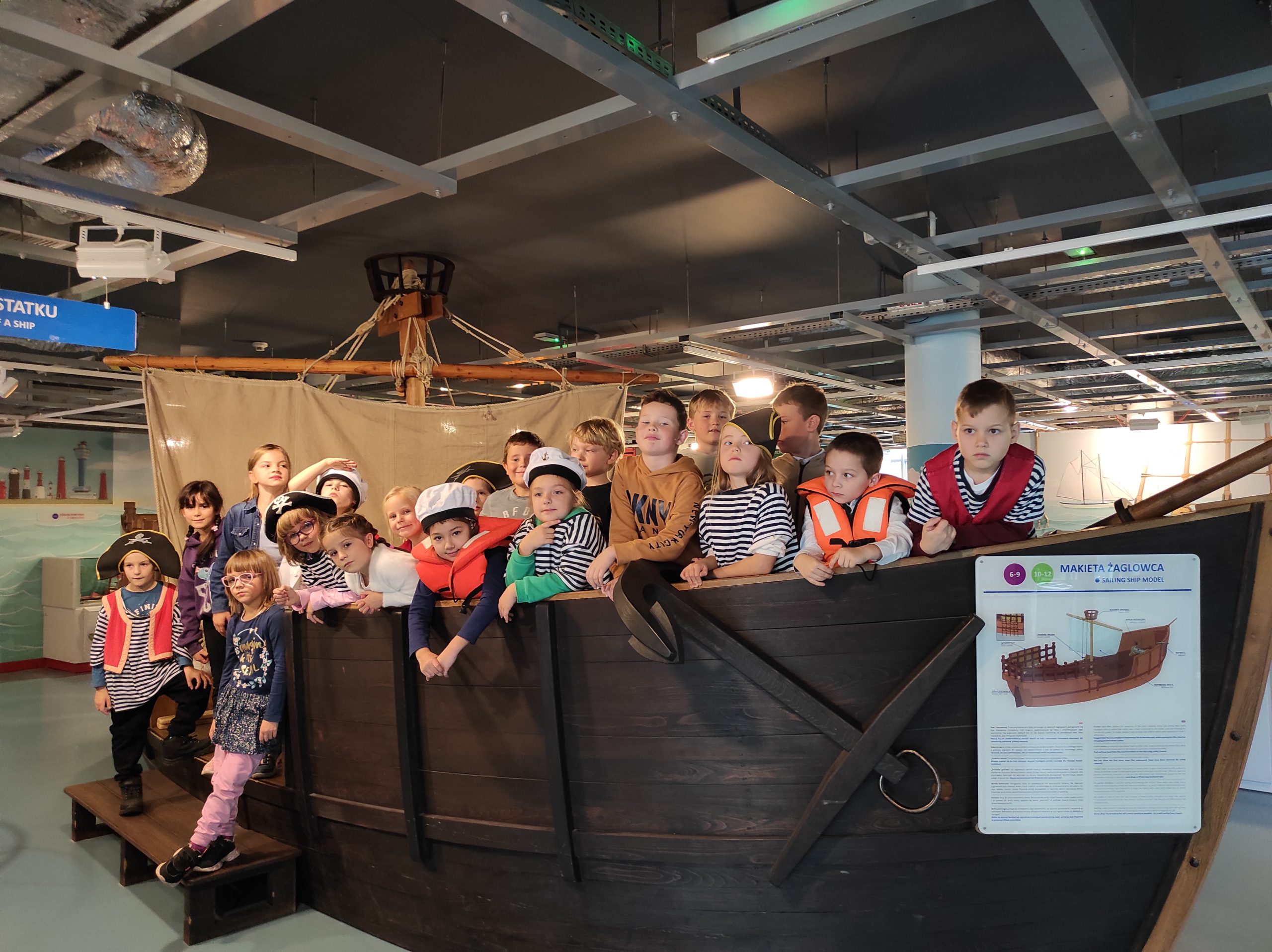 Uczniowie ustawieni w żaglowcu w dwóch rzędach, dwoje dzieci poniżej, stoi na schodku. Niektórzy uczniowie mają na sobie stroje piratów, marynarzy. W tle wystrój Ośrodka Kultury Morskiej.