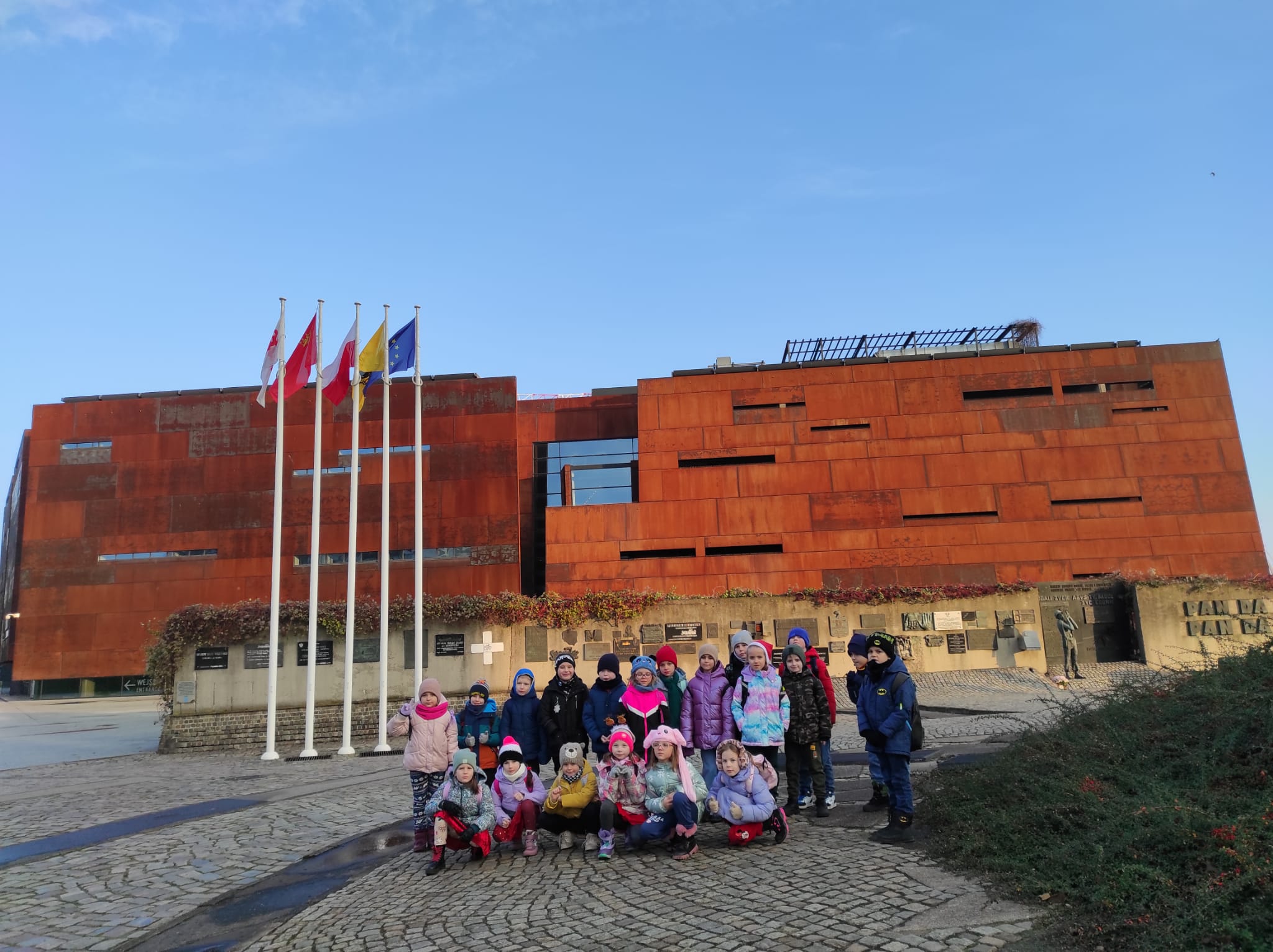 Dzieci ustawione w dwóch rzędach, jeden stoi, drugi siedzi. W tle budynek Europejskiego Centrum Solidarności.