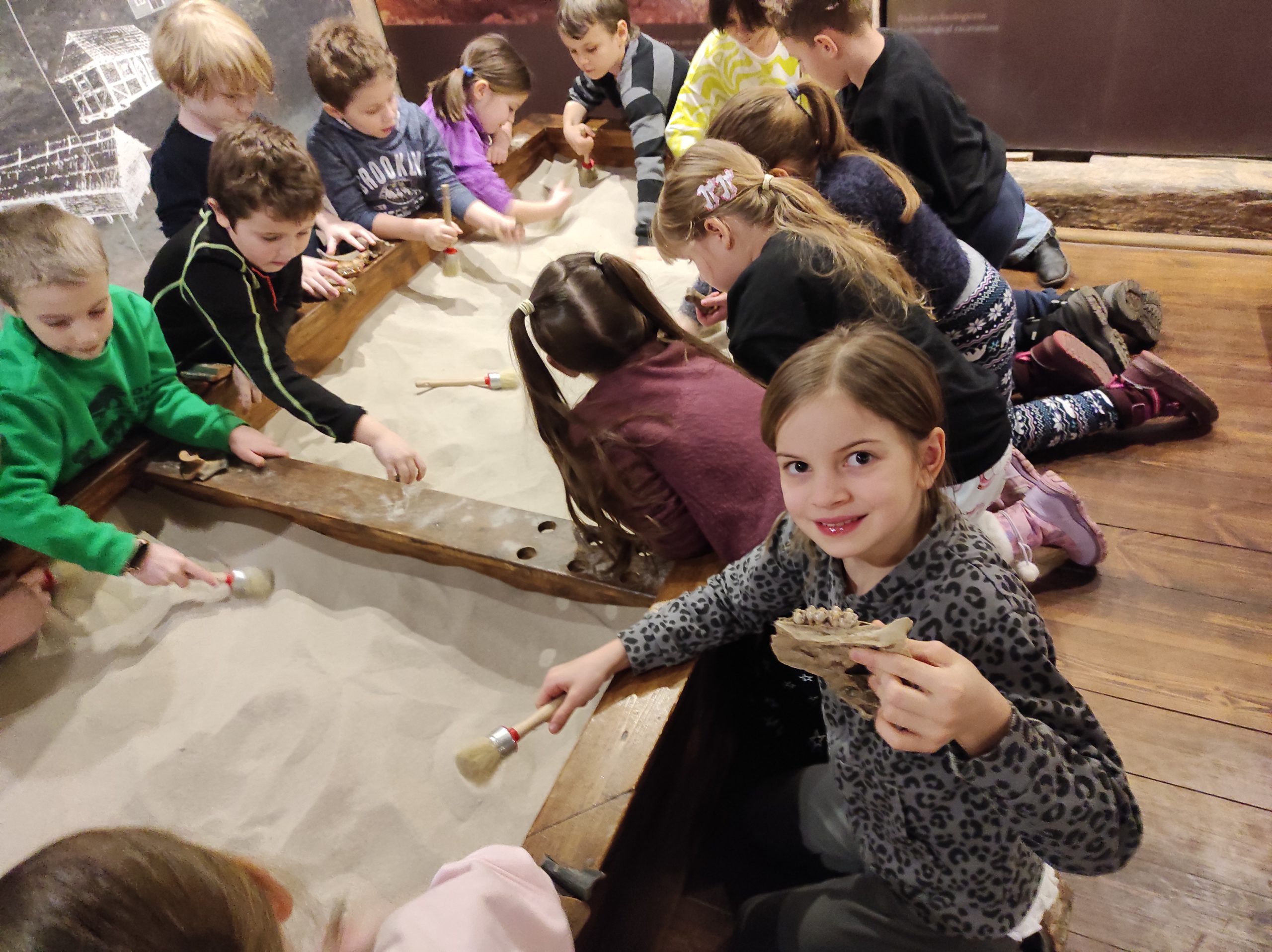 4 dzieci usadzonych wokół piaskownicy. Wszyscy trzymają w dłoniach pędzle. Jedna dziewczynka pozuje do zdjęcia i trzyma w dłoni znalezisko przypominające kawałek kości.