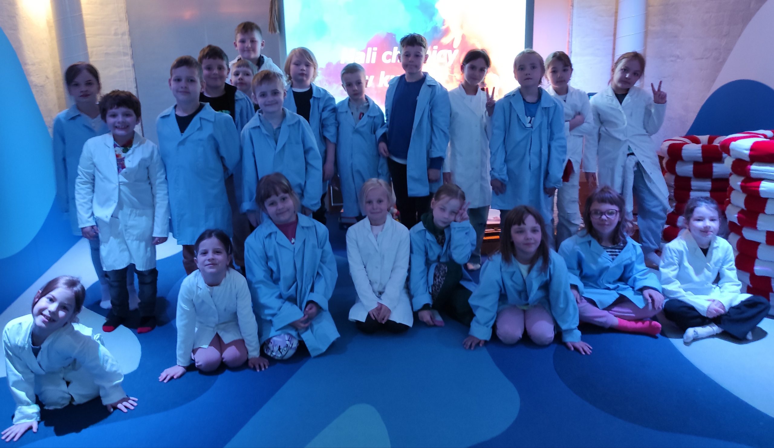 Dzieci ustawione w dwóch rzędach, 1 stoi, 2 siedzi na podłodze. Wszyscy mają ubrane fartuchy. W tle podświetlony ekran oraz ściana.