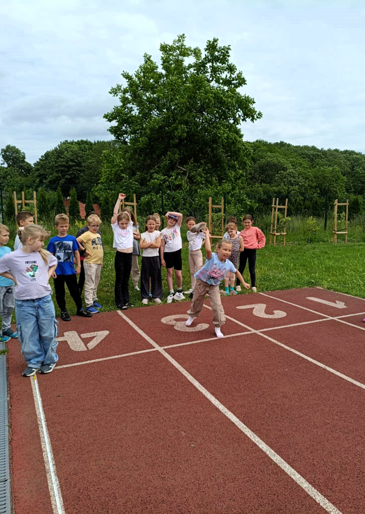 Na pierwszym planie dziewczynka rzuca butem. Za nią stoi grupa dzieci 