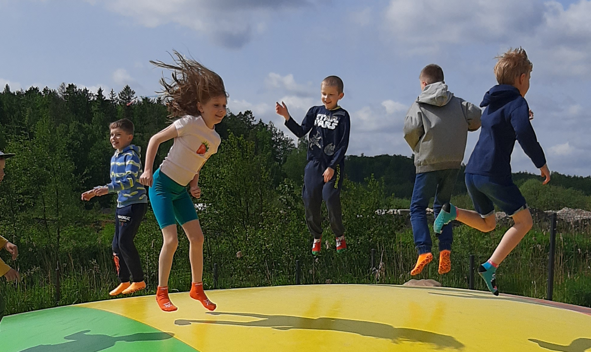 Zdjęcie w plenerze. Grupa 6 dzieci skacze po dmuchańcu. Czworo z nich unosi się w powietrzu. Ich włosy rozwiewa wiatr. W tle pas zieleni. Na niebie pojedyncze chmury.