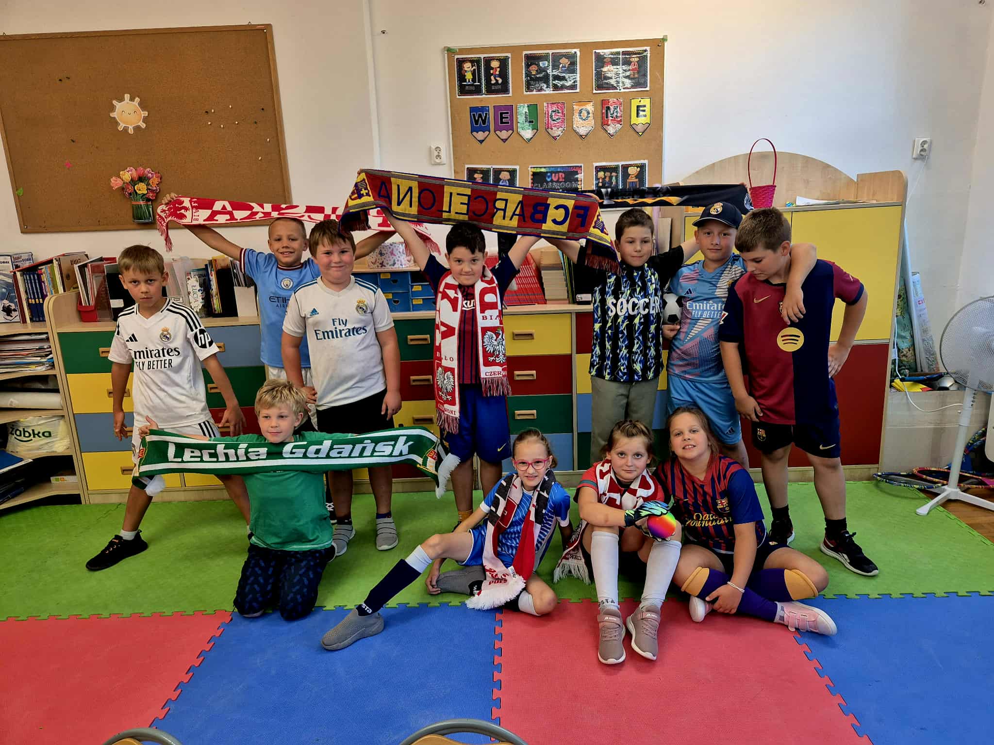 Jedenaście dzieci stoi. Dzieci są w strojach sportowych w strojach piłkarskich. A na Macie siedzą3 dziewczynki. Czworo chłopców stojących trzyma w rękach rozciągnięte szaliki drużyn piłkarskich. Jeden chłopiec stoi w rozkroku. Za dziećmi znajdują się półki na książki i tablica korkowa