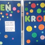 Na dwóch niebieskich tablicach jest duży, kolorowy napis "Dzień Kropki". Po lewej stronie  jest postać dziewczynki z pędzlem, która maluje różnej wielkości, kolorowe kropki, a obok niej napisy: ' Od maleńskiej kropki  wszystko sie zaczyna" i  " Była sobie kropka mała, która talenty odkrywała".