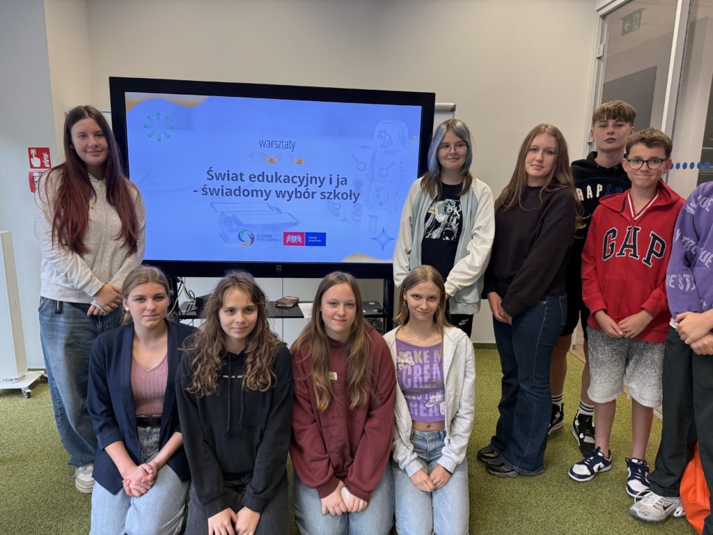 Dziewięcioro uczniów z klasy 8b pozują do zdjęcia podczas warsztatów edukacyjnych pod ha-słem „Świat edukacyjny i ja – świadomy wybór szkoły”. Za nimi widoczny jest ekran z prezentacją.