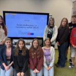 Dziewięcioro uczniów z klasy 8b pozują do zdjęcia podczas warsztatów edukacyjnych pod ha-słem „Świat edukacyjny i ja – świadomy wybór szkoły”. Za nimi widoczny jest ekran z prezentacją.