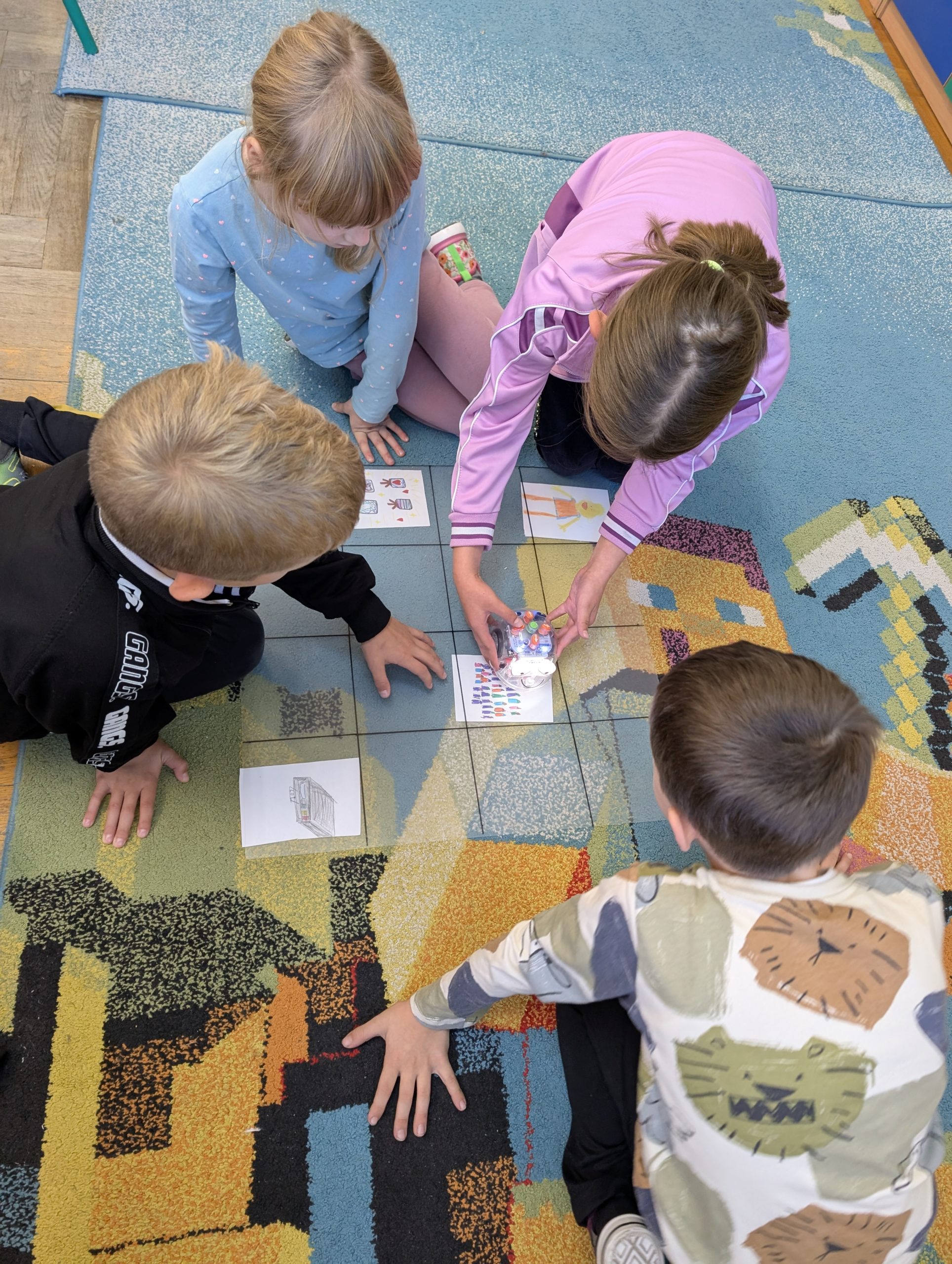 2 uczennice i 2 uczniów pochyleni nad przezroczystą planszą w kratkę, pod której leżą w niektórych miejscach rysunki dzieci. Dziewczynka po prawej trzyma dłoń na blue bocie. Pozostali patrzą na robota