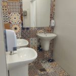 Łazienka z kolorowymi, wzorzystymi płytkami w stylu marokańskim na podłodze i ścianie wokół lustra. Znajdują się tu trzy białe umywalki, duże lustro, dozowniki na mydło i suszarka do rąk.