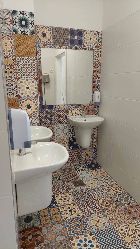 Łazienka z kolorowymi, wzorzystymi płytkami w stylu marokańskim na podłodze i ścianie wokół lustra. Znajdują się tu trzy białe umywalki, duże lustro, dozowniki na mydło i suszarka do rąk.