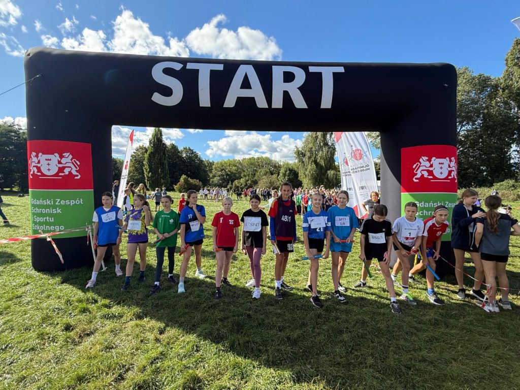 Grupa młodych zawodniczek stoi na linii startu pod dużą czarną bramą z napisem START pod-czas zawodów biegowych na świeżym powietrzu. W tle widoczna jest zieleń parku, kibice oraz ba-nery organizatora – Gdańskiego Zespołu Schronisk i Sportu Szkolnego.