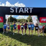 Grupa młodych zawodniczek stoi na linii startu pod dużą czarną bramą z napisem START pod-czas zawodów biegowych na świeżym powietrzu. W tle widoczna jest zieleń parku, kibice oraz ba-nery organizatora – Gdańskiego Zespołu Schronisk i Sportu Szkolnego.