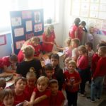 Na półpiętrze szkoły przy stolikach 8 uczniów Koła PP HOPR dokonuje pomiaru ciśnienia uczniom klas młodszych stojących przed stolikami. W tle na tablicy plakaty z sercem.