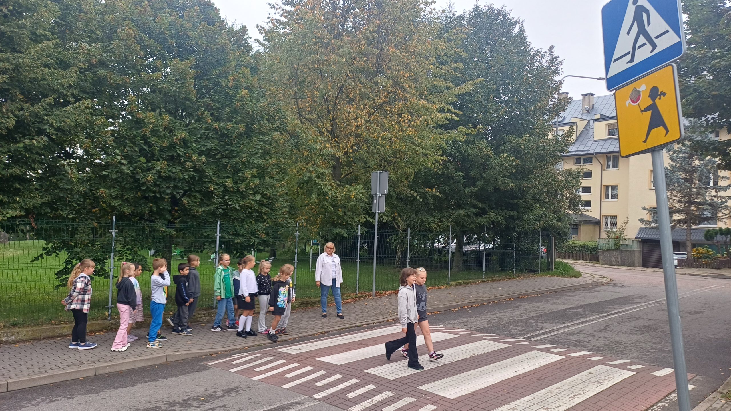 Dwójka dzieci przechodzi przez przejście dla pieszych. Grupa 17 dzieci wraz z nauczycielem stoi na chodniku.