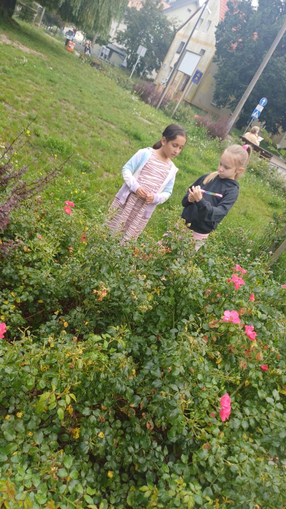 2 dziewczynki stoją przed krzewem róż. Jedna z nich trzyma w rękach notes i długopis.