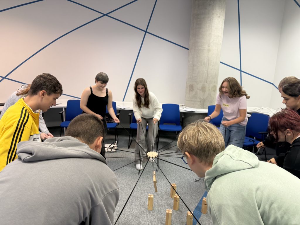 Uczniowie klasy 8b stoją w kręgu i biorą udział w ćwiczeniu integracyjnym. Każdy z nich trzyma sznurek połączony ze wspólną konstrukcją, która umożliwia przenoszenie drewnianych klocków. Uczestnicy współpracują, aby wspólnie ułożyć elementy. W tle znajdują się niebieskie krzesła ustawione przy ścianie ozdobionej geometrycznymi liniami.