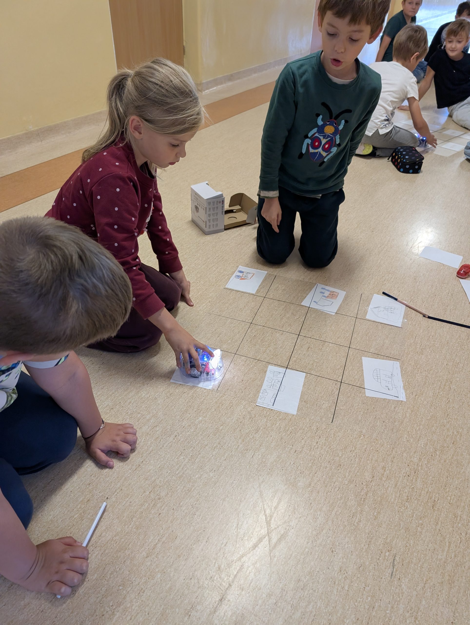 Uczniowie pochyleni nad przezroczystą planszą w kratkę, pod której leżą w niektórych miejscach rysunki dzieci. Dziewczynka po lewej trzyma dłoń na blue bocie. Dwóch chłopców patrzy na robota. 