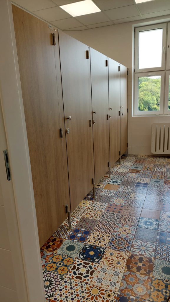 Toaleta z drewnianymi drzwiami kabin i kolorową, mozaikową podłogą w orientalne wzory. Jasne, dobrze oświetlone wnętrze z dużym oknem i widokiem na zieleń.