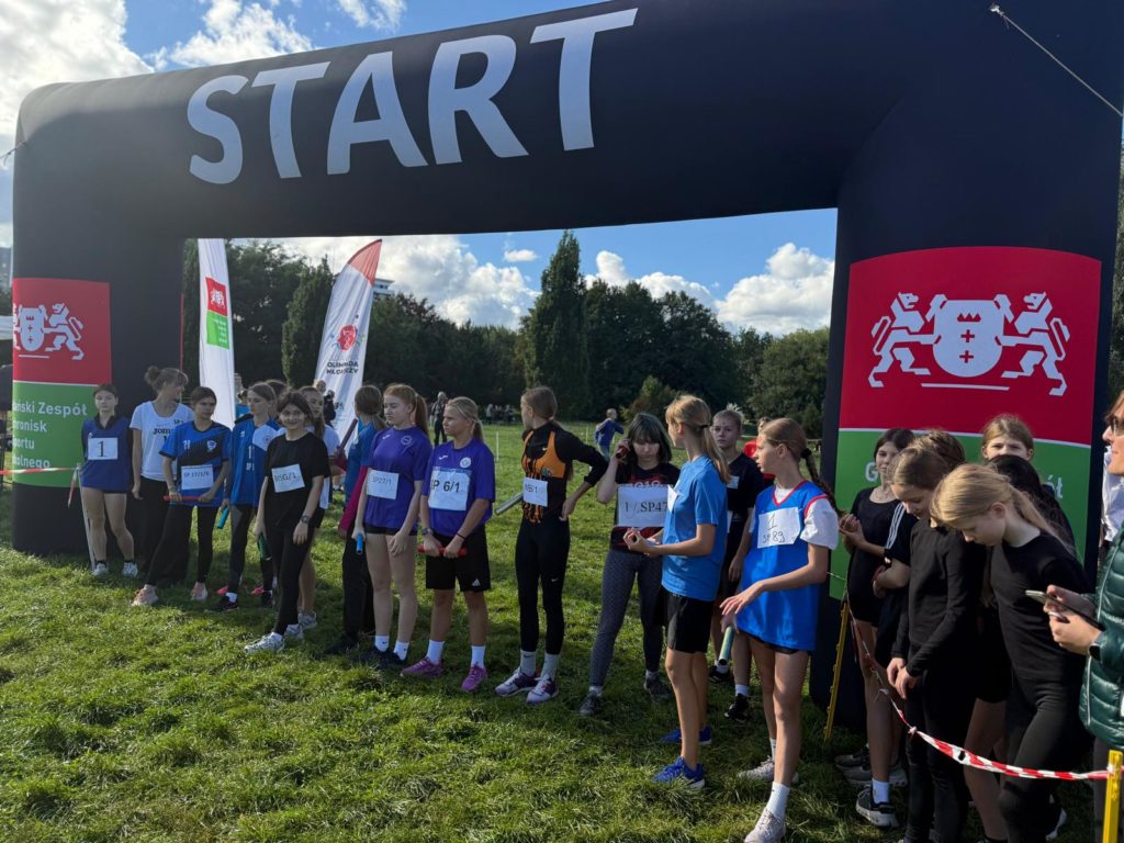 Grupa dziewcząt w sportowych strojach stoi na linii startu pod czarną bramą z napisem START podczas zawodów biegowych w plenerze. Zawodniczki trzymają pałeczki sztafetowe, a w tle wi-doczne są banery Gdańskiego Zespołu Schronisk i Sportu Szkolnego oraz zielone drzewa.