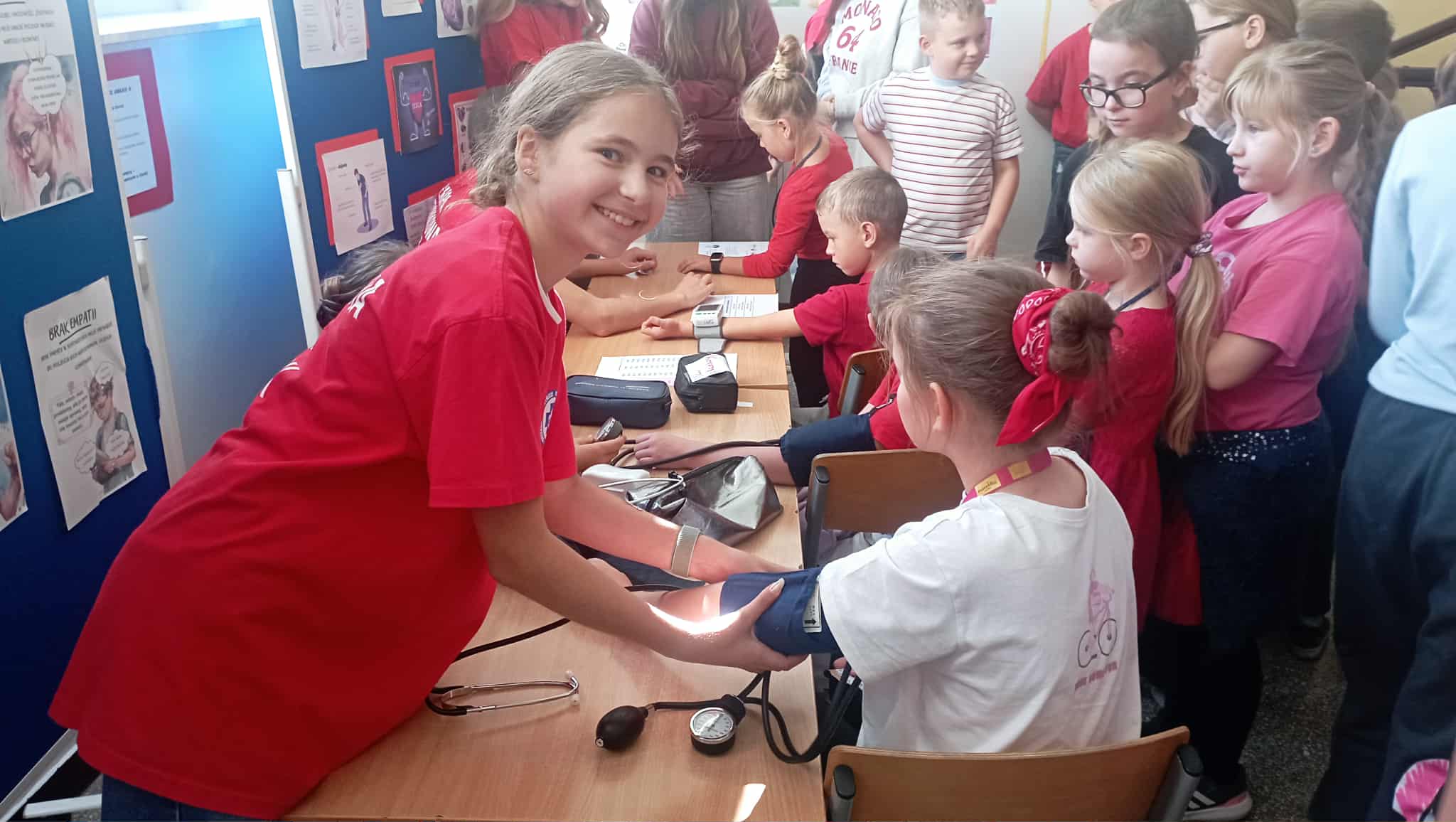 Na pierwszym planie uczennica klasy 5 z Koła PP HOPR mierząca ciśnienie krwi swojej koleżnce. W koło stolika inni uczniowie.