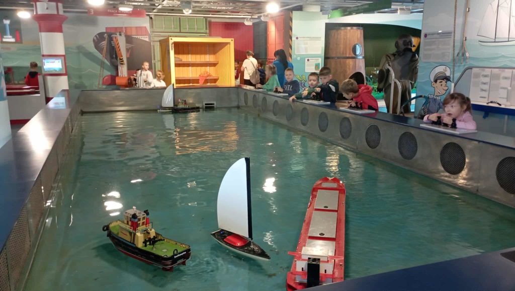 Na pierwszym planie basen z wodą, po którym pływają statki: żaglówki, kontenerowiec i łódź ratunkowa, sterowane przez 6 uczniów klasy 1c. W tle inni uczniowie przy terminalu kontenerowym.