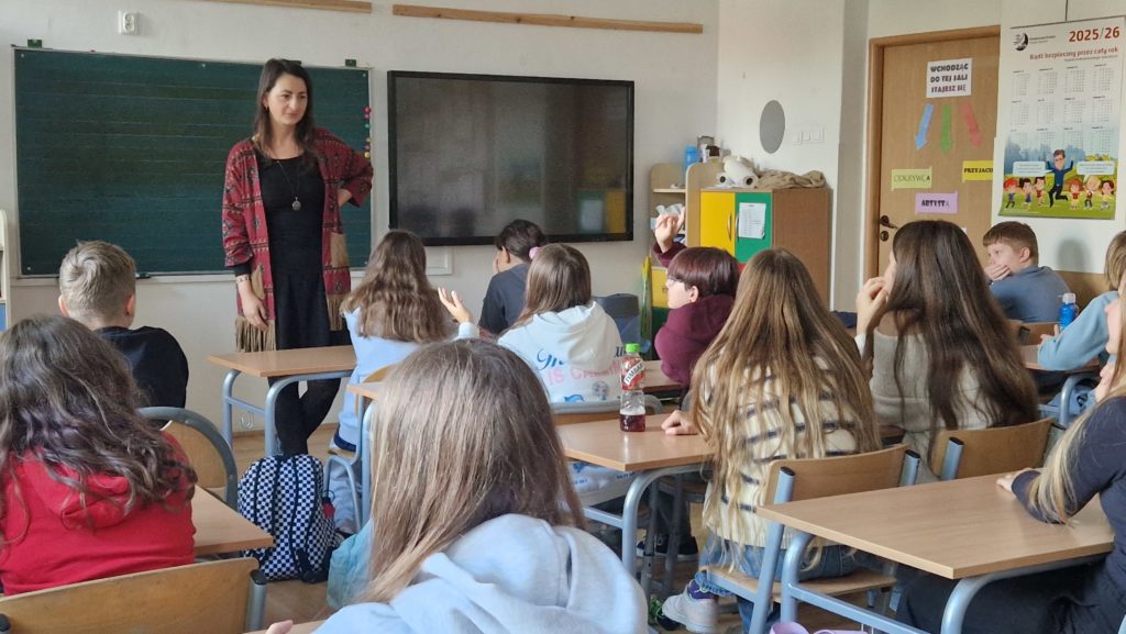 W sali lekcyjnej. Nauczycielka stoi z przodu klasy obok zielonej tablicy i ciemnego monitora lub tablicy interaktywnej. Ubrana jest w czarną sukienkę i czerwony sweter z wzorami. Wokół niej siedzi kilkanaścioro uczniów – większość z nich patrzy na nauczycielkę, a kilku z nich ma podniesione ręce,