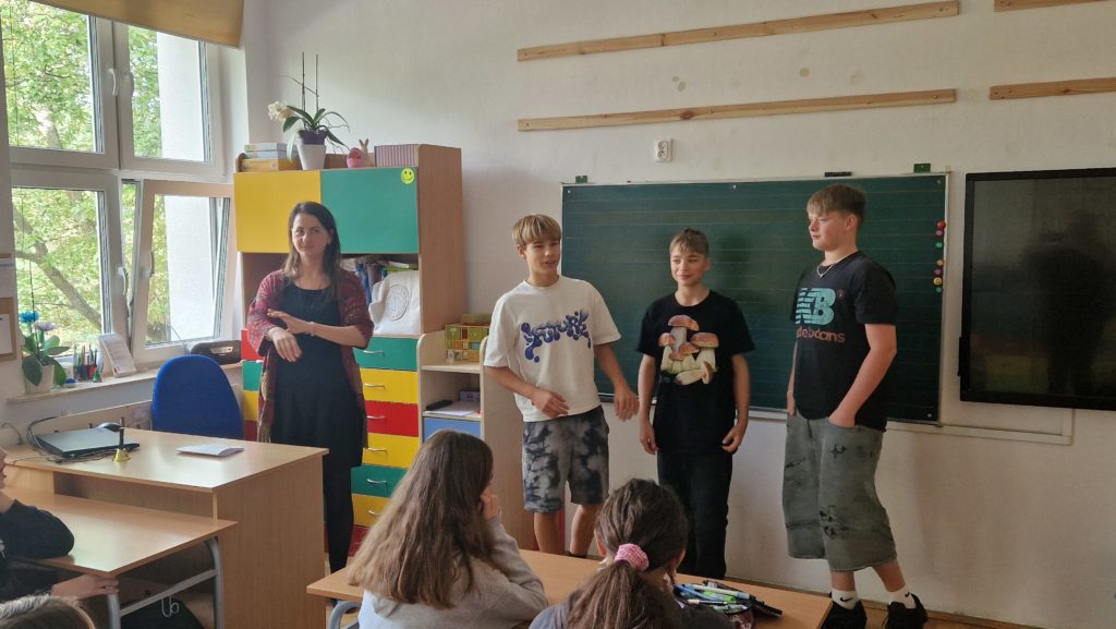 Trzech chłopców stoi obok siebie przodem do klasy, a obok nich nauczycielka, która obserwuje sytuację z uśmiechem. W tle znajduje się zielona tablica i ekran multimedialny. Po lewej stronie widoczna jest kolorowa szafka oraz biurko nauczyciela z laptopem i dzwonkiem. Kilkoro uczniów siedzi w ławkach przodem do prezentujących.