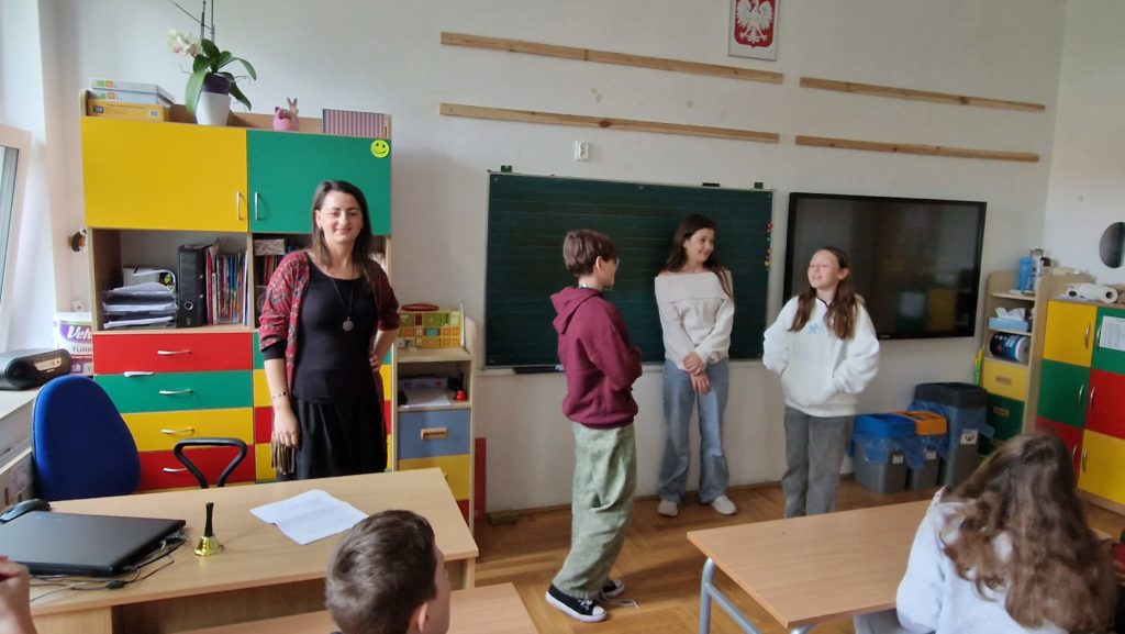 W szkolnej klasie trzy dziewczynki – stoją przy zielonej tablicy, biorąc udział w psychodramie. Obok nich stoi nauczycielka uśmiechnięta i obserwująca uczniów. Pozostali uczniowie siedzą w ławkach, zwróceni w stronę tablicy.