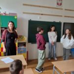 W szkolnej klasie trzy dziewczynki – stoją przy zielonej tablicy, biorąc udział w psychodramie. Obok nich stoi nauczycielka uśmiechnięta i obserwująca uczniów. Pozostali uczniowie siedzą w ławkach, zwróceni w stronę tablicy.