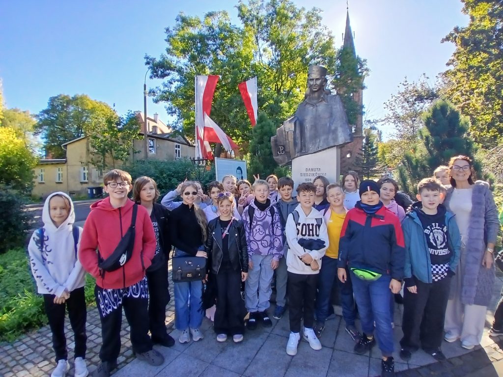 Grupa dzieci z nauczycielką pozuje przed pomnikiem Danuty Siedzikówny „Inki”. W tle widać flagi Polski, kościół i drzewa. Zdjęcie zrobiono podczas słonecznego dnia.