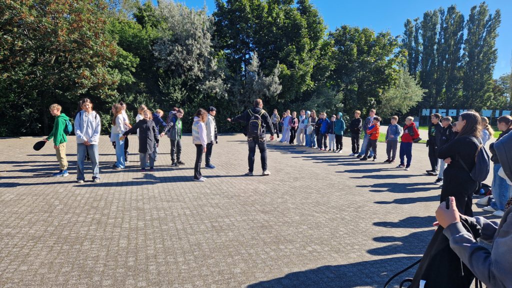 Grupa młodzieży stoi w okręgu na świeżym powietrzu, uczestnicząc w zajęciach prowadzonych przez dorosłego. W tle widoczny jest fragment napisu „NIGDY WIĘCEJ WOJNY”, sugerujący miejsce pamięci.