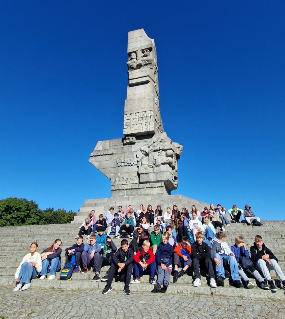 Grupa młodych osób siedzących na schodach pomnika. W tle znajduje się monumentalna budowla z kamienia — to Pomnik Obrońców Wybrzeża na Westerplatte w Gdańsku.