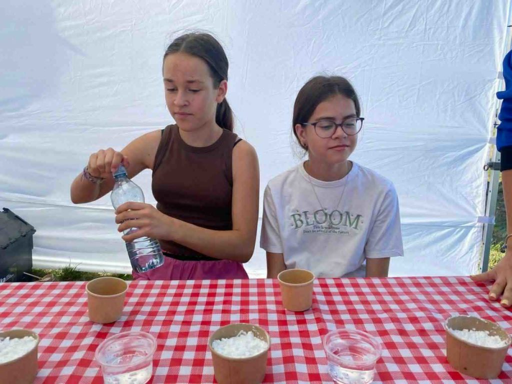 Dwie uczennice siedzą przy stole, na którym znajdują się brązowe i przezroczyste kubki