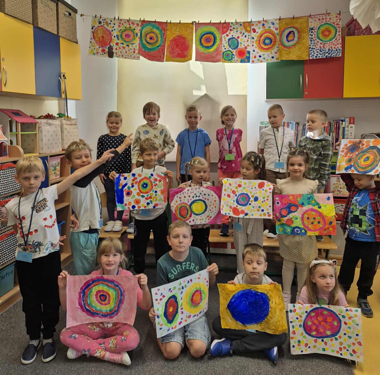  Dzieci ustawione w trzech rzędach  w świetlicy trzymają w dłoniach  swoje kolorowe prace  przedstawiające różnej wielkości kropki. W tle znajdują się  kolorowe szafki. Nad dziećmi są kolejne prace o kropkach przyczepione klamerkami do sznurka.