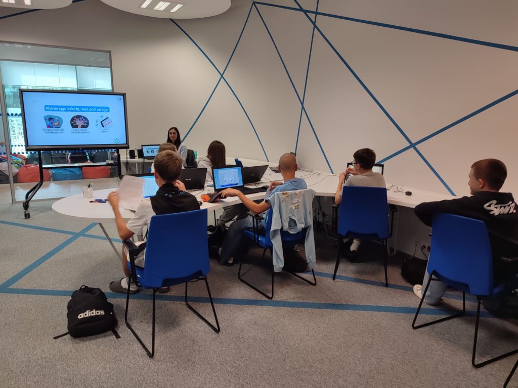 Uczniowie z klasy 8a siedzą przy biurkach i pracują z laptopami. Przed nimi stoi duży ekran, na którym wyświetlana jest prezentacja. Zajęcia prowadzi osoba siedząca przy biurku obok ekranu. Pomieszczenie jest nowoczesne, z białymi ścianami ozdobionymi niebieskimi liniami i wygodnymi, niebieskimi krzesłami.