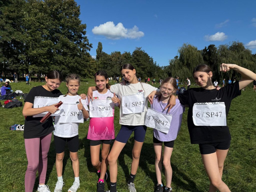 Sześć dziewcząt z numerami startowymi SP47 pozuje razem na trawie podczas zawodów biego-wych w plenerze. Uśmiechnięte zawodniczki stoją w rzędzie, obejmują się i prezentują sportowe stroje, a jedna z nich trzyma pałeczkę sztafetową. W tle widoczny jest park i inni uczestnicy wyda-rzenia.