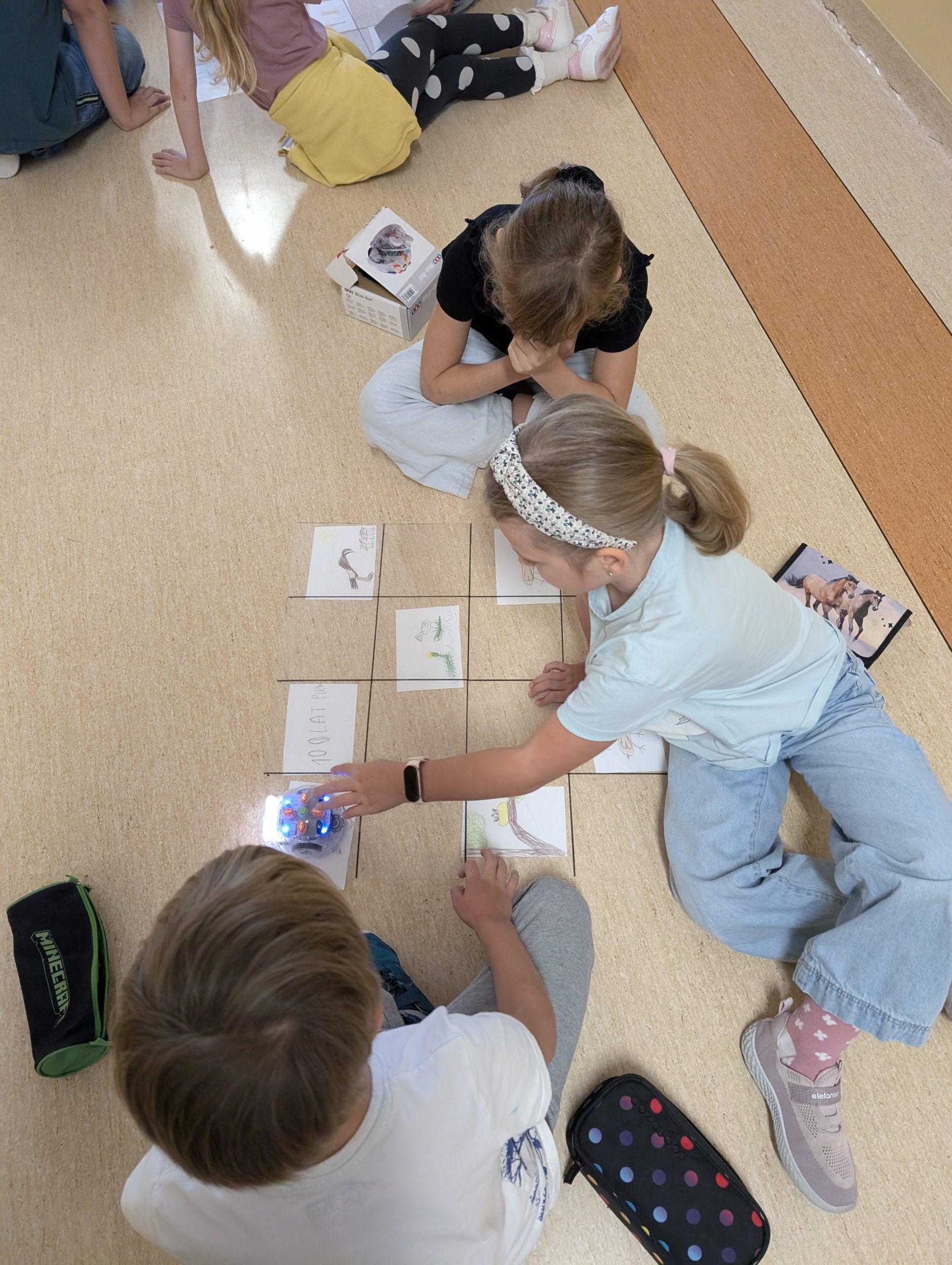 Uczniowie pochyleni nad przezroczystą planszą w kratkę, pod której leżą w niektórych miejscach rysunki dzieci. Dziewczynka po prawej trzyma dłoń na blue bocie. Druga dziewczynka i chłopiec patrzą na robota. 
