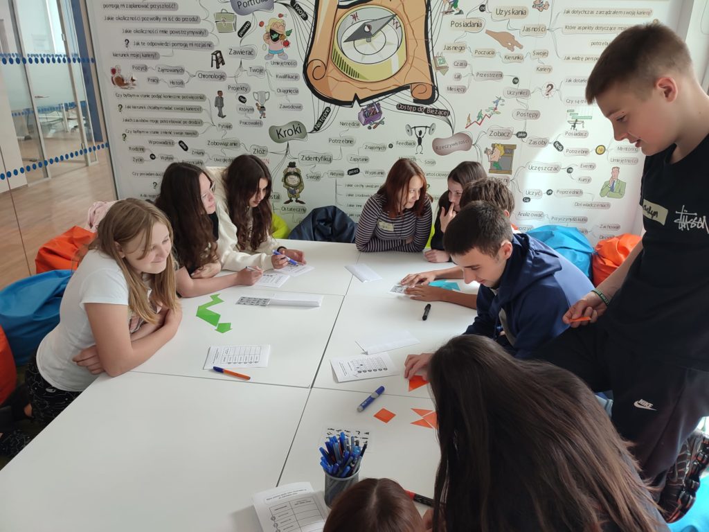 Uczniowie z klasy 8a siedzą wspólnie przy dużym białym stole, rozwiązując zadania i układając kolorowe figury geometryczne. W tle widoczna jest ściana z kolorową grafiką i hasłami edukacyj-nymi. Uczniowie uśmiechają się i współpracują.