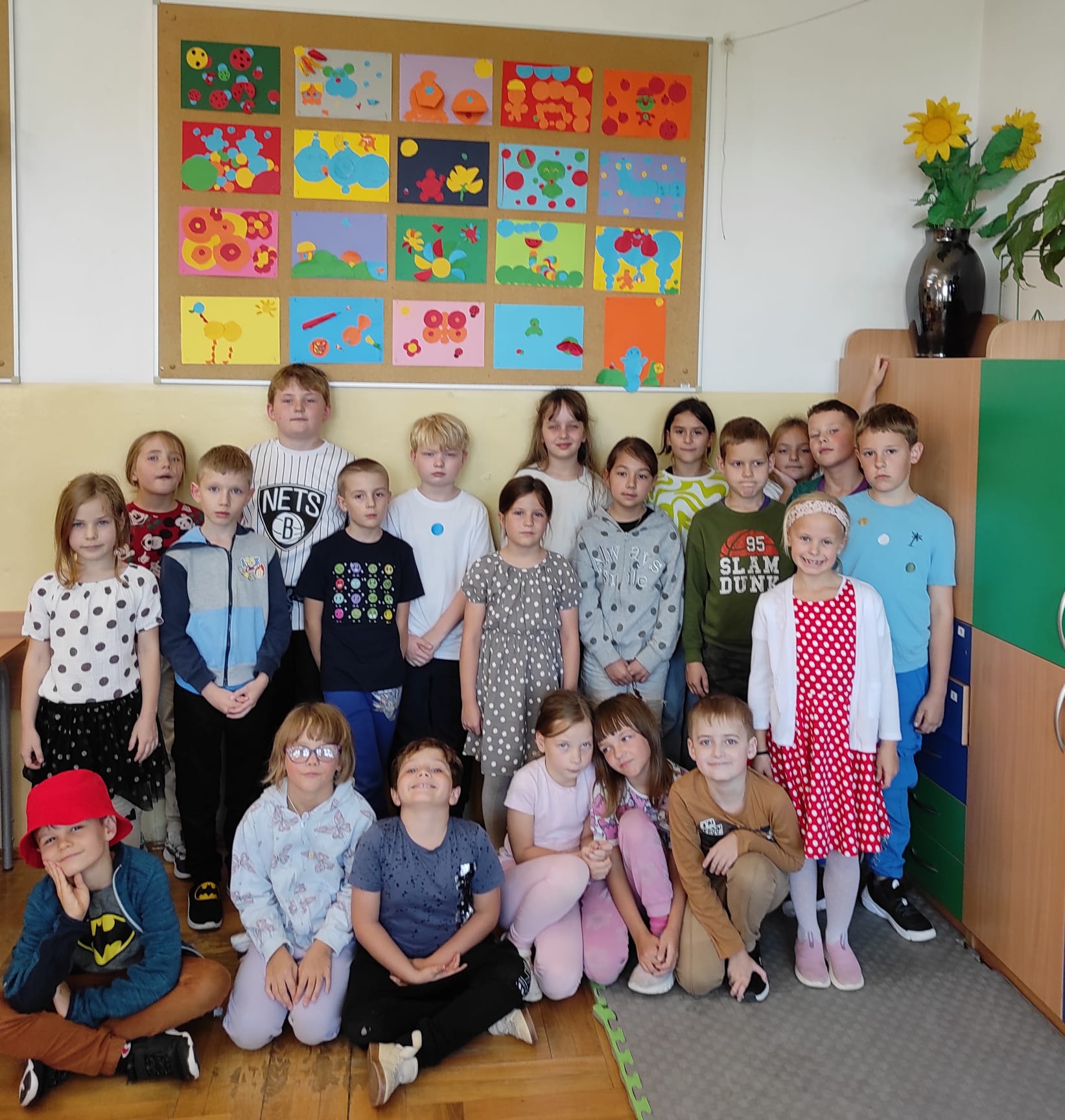 Dzieci ustawione w trzech rzędach w sali lekcyjnej pozują na tle tablicy, na której  wyeksponowane  są ich prace plastyczne z kółek origami. Większość dzieci ma na sobie ubranie w kropki.