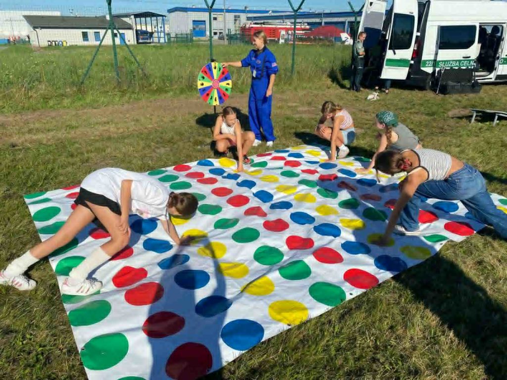 Na trawie leży plansza do gry w twistera, na której znajdują się 4 dziewczynki, obok stoi prowadząca która prowadzi grę 